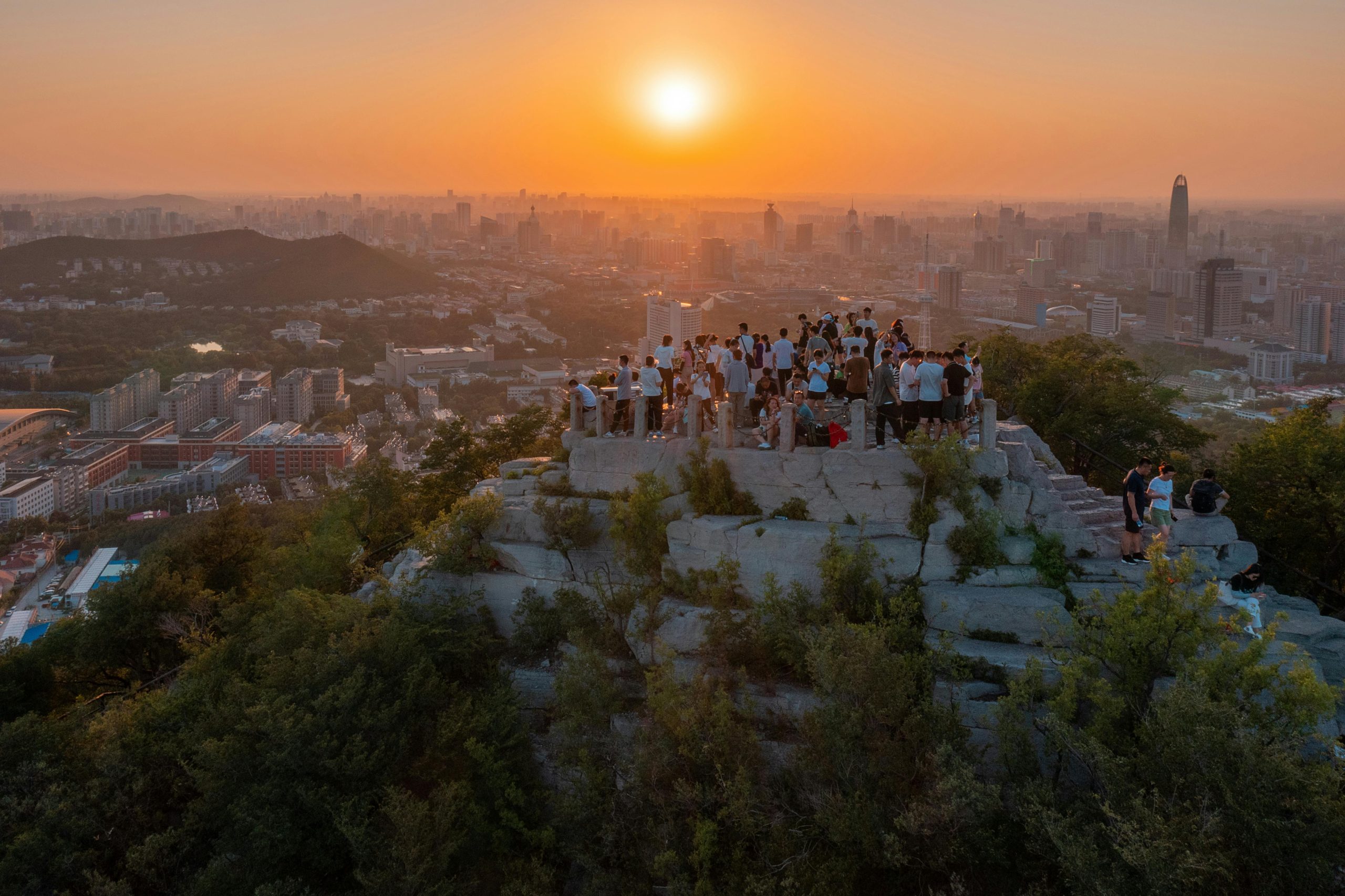 Breathtaking sunset view over Jinan city skyline from a rocky hilltop with a crowd enjoying the urban scenery.