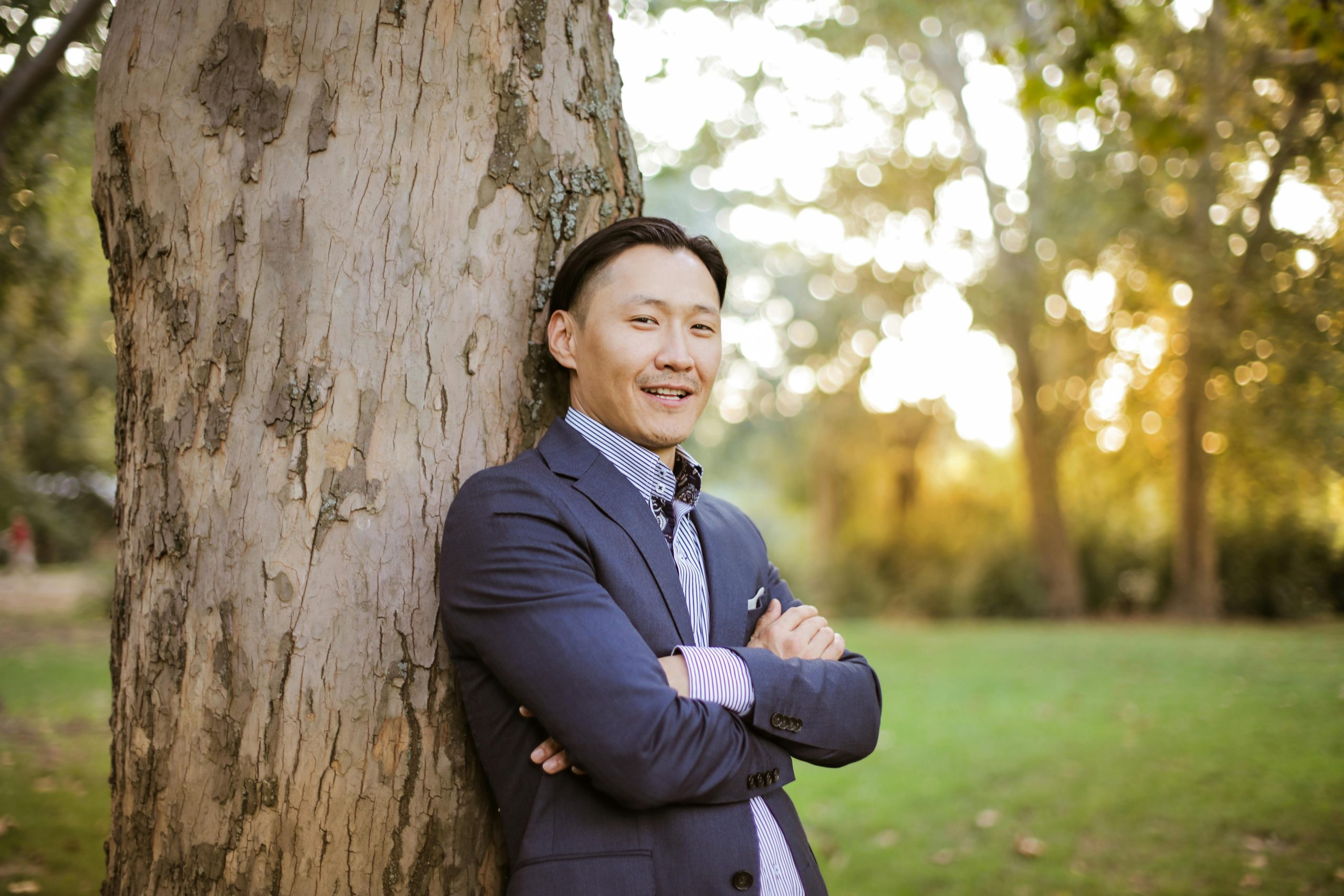 A confident man in formal attire smiling and leaning against a tree outdoors.