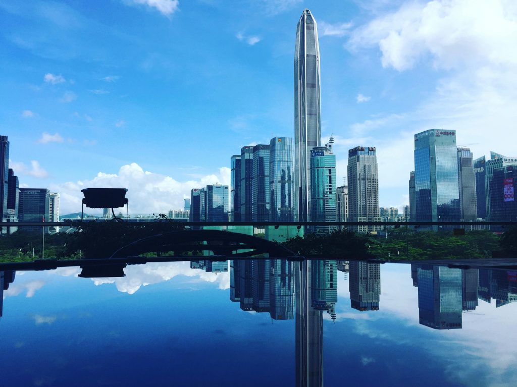 Urban skyline featuring skyscrapers and reflections in a river under a clear blue sky.